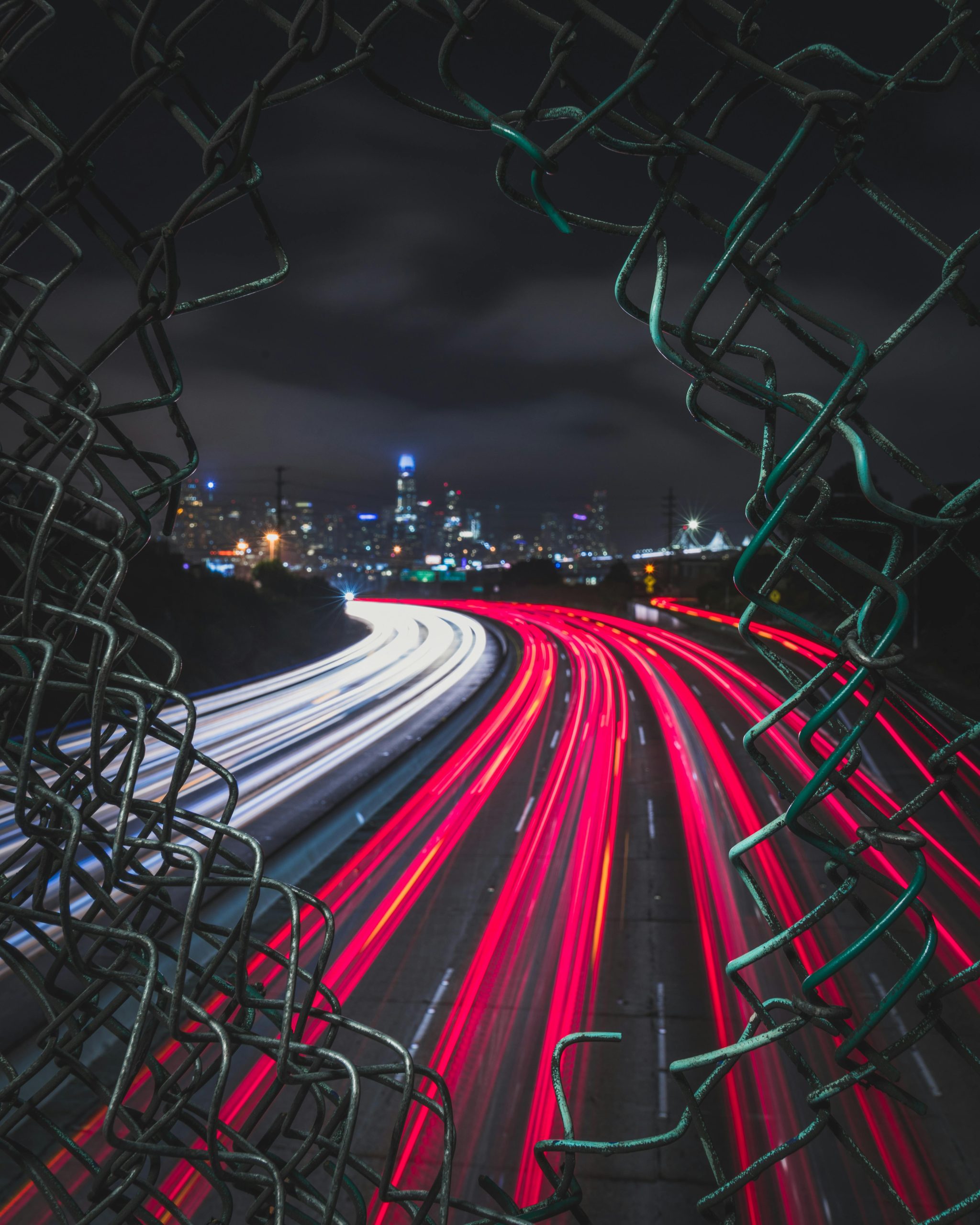 Long exposure captures urban expressway lights through a broken fence at night in San Francisco.